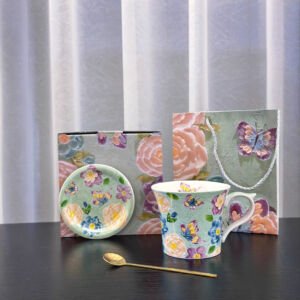 A ceramic mug and saucer set arranged on a surface, featuring a green background decorated with colorful butterflies and floral patterns, accompanied by a gold spoon, a matching box, and a gift bag