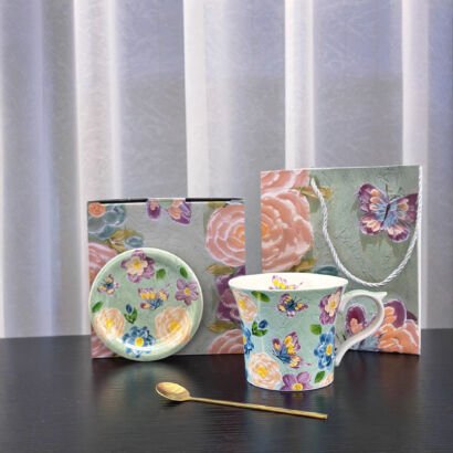 A ceramic mug and saucer set arranged on a surface, featuring a green background decorated with colorful butterflies and floral patterns, accompanied by a gold spoon, a matching box, and a gift bag