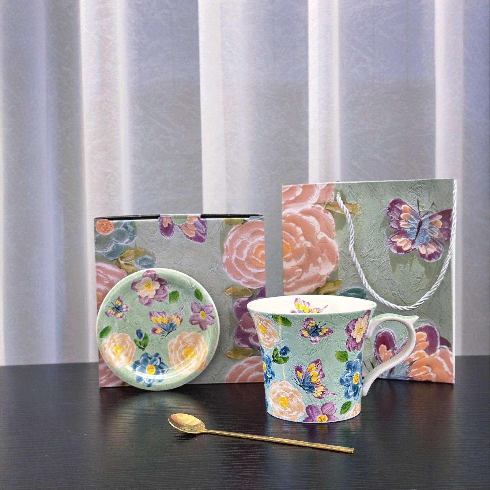 A ceramic mug and saucer set arranged on a surface, featuring a green background decorated with colorful butterflies and floral patterns, accompanied by a gold spoon, a matching box, and a gift bag