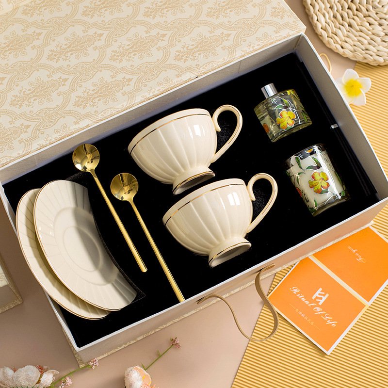 An overhead view of a luxury ceramic teacup and saucer set packaged inside a decorative gift box, featuring two cream-colored fluted teacups, matching saucers, two gold spoons, and two small decorative items including a glass perfume bottle and a glass cup
