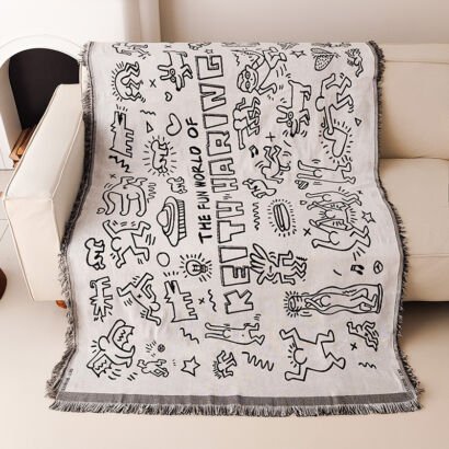 A light-colored woven throw blanket draped over a sofa, featuring black line-art graffiti illustrations in the style of Keith Haring with figures, dogs, and the text "THE FUN WORLD OF KEITH HARING", finished with fringed edges