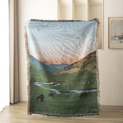 A woven throw blanket hanging indoors, featuring a landscape illustration of horses grazing in a lush green valley with a winding stream, rolling hills, and mountains under a soft sky, finished with fringed edges