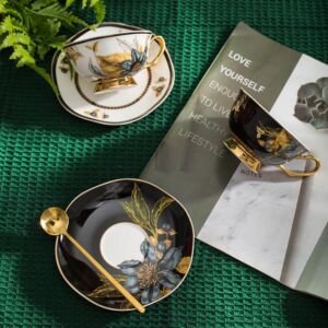 A ceramic teacup and saucer set arranged on a green textured surface, featuring a black set and a white set adorned with metallic gold floral patterns, a gold spoon, and an open lifestyle magazine in the background