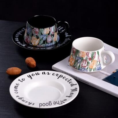 A ceramic teacup and saucer set displayed on a dark surface, featuring two color variations—one black and one white—decorated with a colorful floral tulip pattern, with the inspiring text "May the good things come to you as expected" written along the rim of the saucers