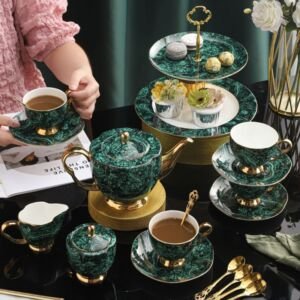 Luxury deep emerald green marbled porcelain tea set arranged on a dark glass table, including a gold-accented teapot, milk pitcher, sugar bowl, teacups with saucers, gold teaspoons, and a matching two-tiered dessert stand filled with pastries