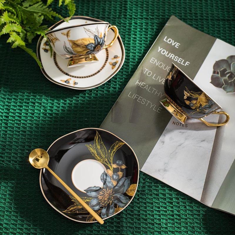 A ceramic teacup and saucer set arranged on a green textured surface, featuring a black set and a white set adorned with metallic gold floral patterns, a gold spoon, and an open lifestyle magazine in the background