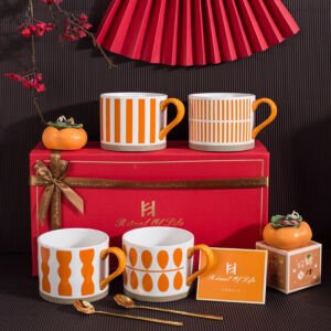 A luxury ceramic mug set arranged on a surface, featuring four white mugs with various orange geometric patterns and handles, two spoons, orange persimmon-shaped ornaments, and a red gift box labeled "Ritual of Life"