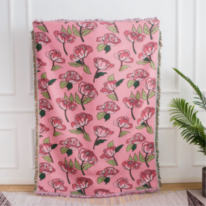 A pink woven throw blanket hanging against a white wall, featuring an all-over repeating pattern of pink roses with green leaves and fringed edges
