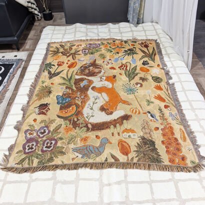 A woven throw blanket spread out on a bed, featuring a detailed vintage-style illustration of a cat and a fox surrounded by flowers, plants, and small animals on a beige background with fringed edges