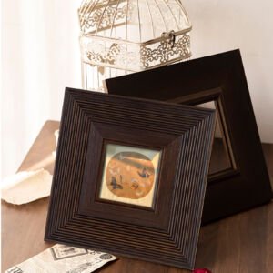 Two dark brown square wooden picture frames leaning on a wooden table, the front frame featuring a stepped ribbed texture and displaying a small circular painting, with a white wire birdcage in the background