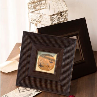 Two dark brown square wooden picture frames leaning on a wooden table, the front frame featuring a stepped ribbed texture and displaying a small circular painting, with a white wire birdcage in the background