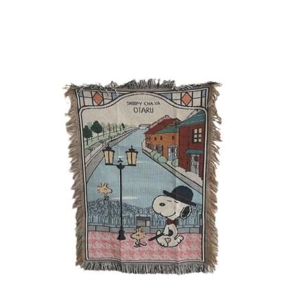A woven throw blanket featuring an illustrated scene of Snoopy wearing a bowler hat and bowtie next to a lamppost overlooking a canal with historic buildings, with the text "SNOOPY CHA-YA OTARU" at the top and fringed edges