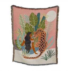 A woven throw blanket displaying a tropical illustration of a woman with long dark hair sitting next to a leopard among lush jungle leaves and a white moon against a pink and cream background, with fringed edges