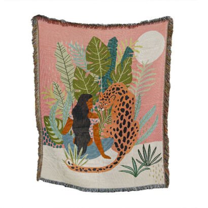 A woven throw blanket displaying a tropical illustration of a woman with long dark hair sitting next to a leopard among lush jungle leaves and a white moon against a pink and cream background, with fringed edges