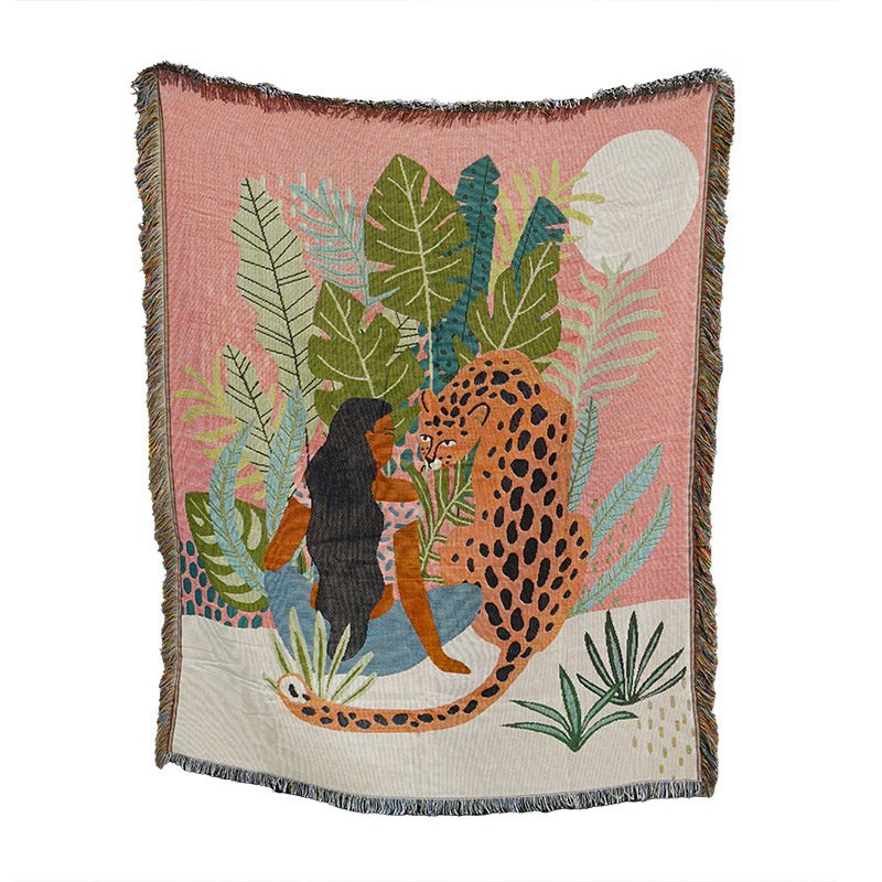 A woven throw blanket displaying a tropical illustration of a woman with long dark hair sitting next to a leopard among lush jungle leaves and a white moon against a pink and cream background, with fringed edges