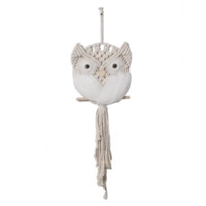 A handcrafted white macrame wall hanging shaped like an owl, featuring intricate woven knots, large round eyes, feathered wings, and a long cascading fringe tail perched on a small wooden stick