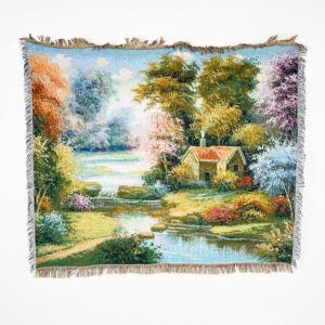 A rectangular woven tapestry throw blanket featuring a picturesque landscape illustration of a cottage by a river, surrounded by lush colorful trees and a flowing stream, with fringed edges