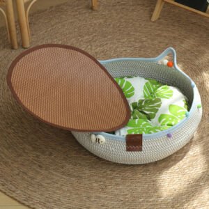 A round, grey and light blue woven cotton rope cat bed shaped with cat ears, containing a soft green monstera leaf print cushion, a cute small fish emblem on the rim, and a removable brown woven bamboo cooling mat set on a jute rug.