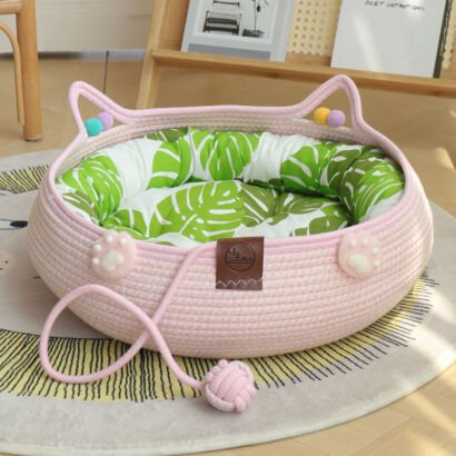 A light pink woven cotton rope cat bed with cute cat-ear handles, plush cat paw accents, colorful pom-poms, an attached pink string ball toy, and a green monstera leaf print inner cushion resting on a round rug.