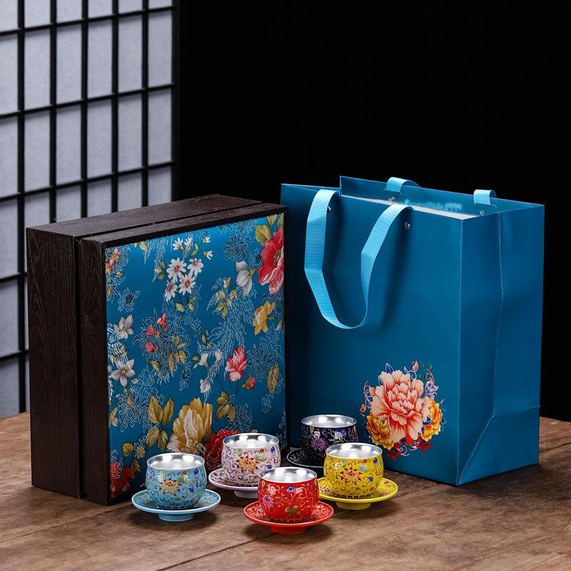 A collection of five multi-colored handcrafted ceramic teacups with silver-lined interiors and matching saucers, arranged in front of a blue floral silk brocade wooden box and a matching blue paper gift bag with peony artwork.