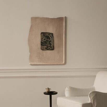 A minimalist interior featuring a large beige textured plaster relief wall sculpture with raw deckled edges and a central dark distressed ceramic patch, hanging on a warm beige wall above a small side table and a white armchair.