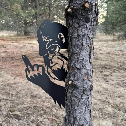 Black metal Bigfoot silhouette tree peeker decor, sneaking sasquatch flipping hand gesture yard art mounted beside pine tree trunk