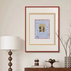 A vertical framed art print hanging above a wooden console table next to a floor lamp, featuring a small central vintage sketch surrounded by intricate blue swirls, a beige inner mat, and a bold red outer frame.