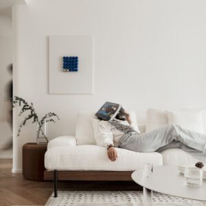 A minimal bright living room featuring a person relaxing on a white modern sofa below a large white canvas framed wall art with a geometric grid of 3D cobalt blue spheres and silver accent balls.