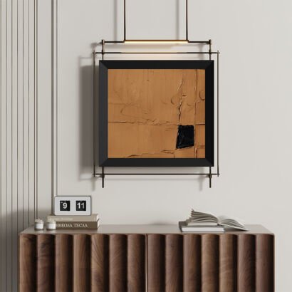 A square artwork with a heavily textured warm ocher and terracotta plaster finish featuring a single black accent block in a thick black frame, suspended within a slim dark metal ceiling-hung frame with integrated LED gallery lighting above a fluted wooden console table.