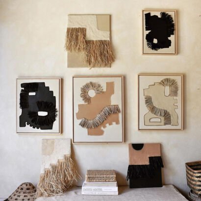 A gallery wall arrangement featuring multi-panel vertical abstract canvas artworks decorated with natural and black raffia straw fringe, layered textured paper, and minimalist earth-tone color blocking in beige, black, and warm terracotta.