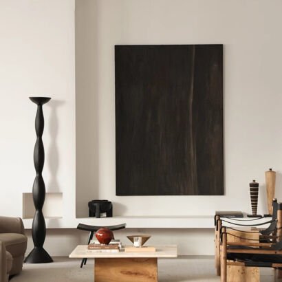A large vertical black and dark espresso brown abstract artwork with subtle wood grain textures mounted on a warm off-white wall in a modern living room with a black floor lamp and low coffee table.