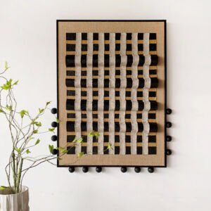 A vertical rectangular artwork in a black frame mounted on a white wall next to a potted plant, featuring a 3D woven grid of textured off-white linen ribbons interwoven with black and natural burlap stripes, accented with black wooden spheres along the frame edges.