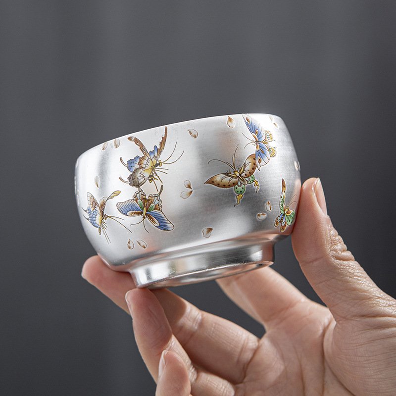 A hand holding a rounded metallic sterling silver teacup with a brushed matte finish, decorated with detailed enamel paintings of colorful fluttering butterflies and delicate falling petals set against a dark background.