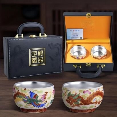 A pair of ceramic teacups with silver-lined interiors and enamel dragon and phoenix motifs shown in the foreground, next to a black leatherette briefcase-style gift box with a yellow padded silk lining and gold hardware.