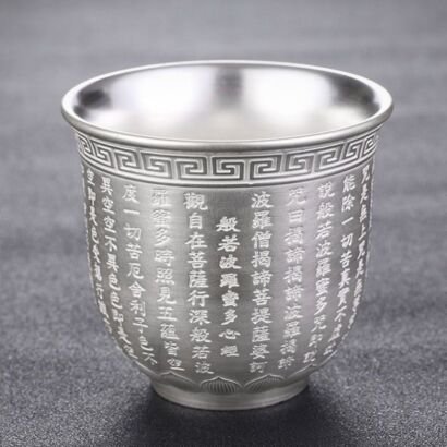 A single metallic silver teacup made of sterling silver, featuring delicate relief-engraved Chinese Heart Sutra characters, a geometric meander pattern along the rim, and a lotus petal motif base set against a dark grey background.
