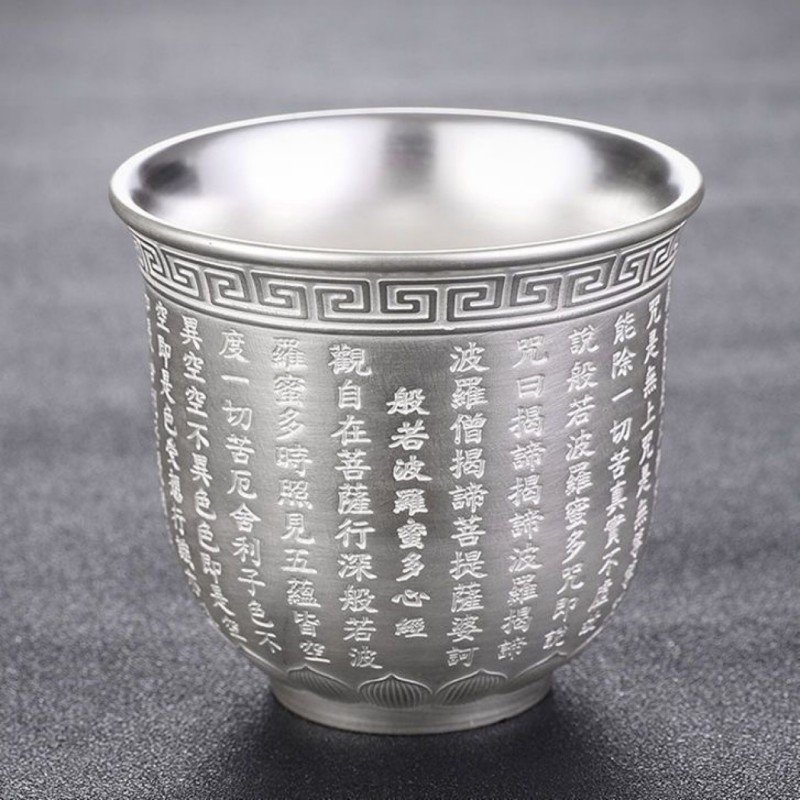 A single metallic silver teacup made of sterling silver, featuring delicate relief-engraved Chinese Heart Sutra characters, a geometric meander pattern along the rim, and a lotus petal motif base set against a dark grey background.
