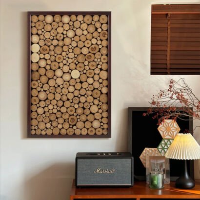 A vertical rectangular artwork in a dark wooden frame filled with closely packed natural wood log slices of varying sizes, mounted on a beige wall above a wooden tabletop console with a vintage speaker and lamp.
