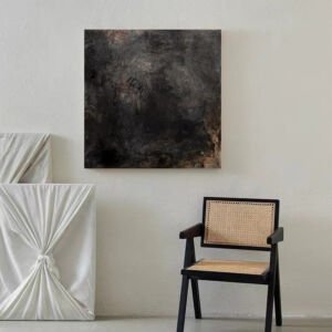 A square abstract canvas artwork with a dark charcoal and moody slate-grey textured concrete surface, hanging on a white textured wall above a modern black wooden armchair with woven rattan seating.