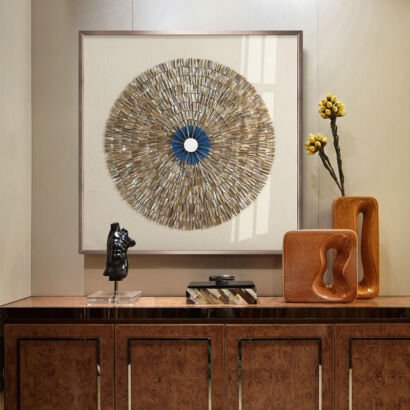 A large square framed wall artwork featuring a circular sunburst mosaic made of iridescent mother-of-pearl tiles with a cobalt blue central ring and mirror core, displayed above a luxury wooden console table decorated with modern sculptures and ceramic vases.