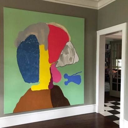 A large square abstract canvas painting featuring bold, oversized color blocking in blue, red, yellow, grey, and brown forming a stylized profile portrait against a sage green background, mounted on a muted grey wall near a doorway.