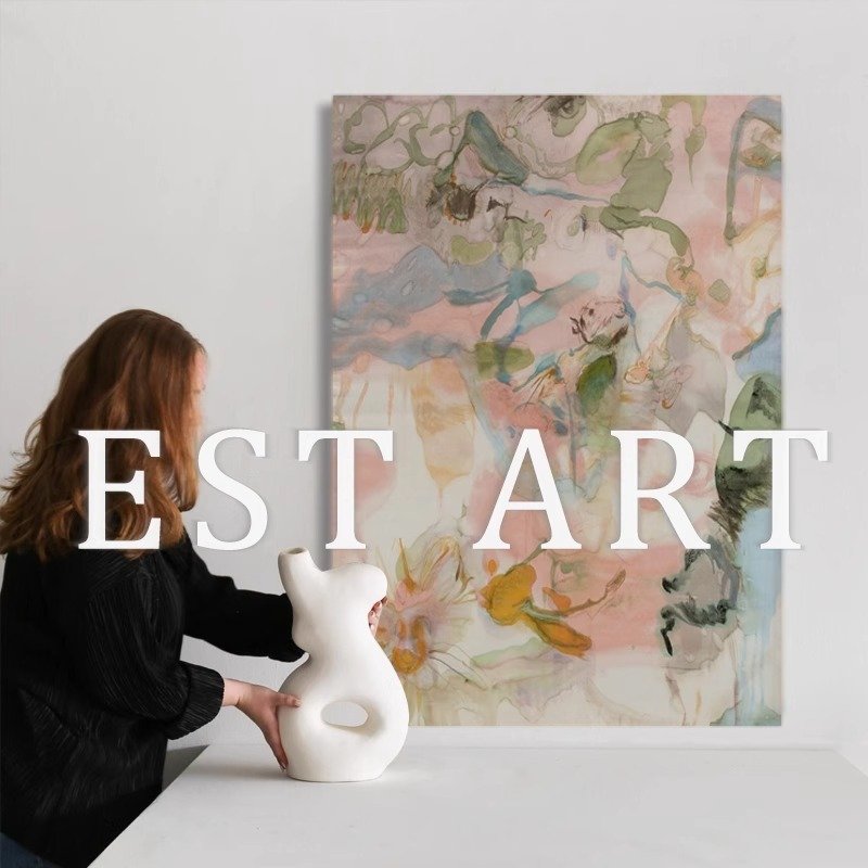 A large vertical framed artwork featuring fluid watercolor abstract brushstrokes in pastel pink, sage green, blush, and golden yellow hues mounted on a white wall, with a woman arranging a modern white ceramic vase in the foreground.