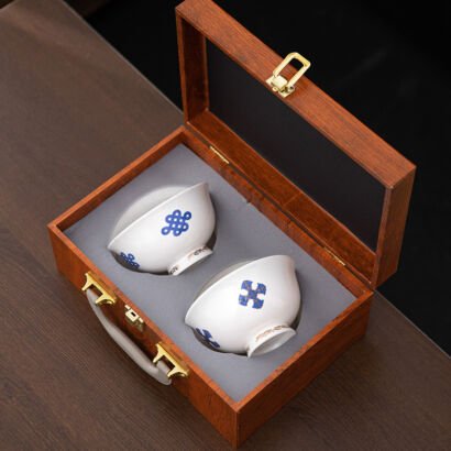An open wooden suitcase-style display box with a glass lid and grey padded interior, revealing two white ceramic teacups featuring blue auspicious knot emblems, gold trim, and silver-lined interiors.