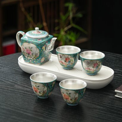 A turquoise floral enamel teapot and four matching ceramic teacups with silver-lined interiors, set on a white zen-patterned tea tray against a dark wooden table.