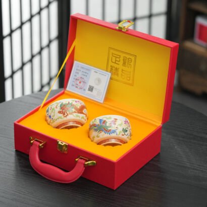 Two handcrafted ceramic teacups featuring silver-lined interiors and colorful Chinese dragon and phoenix enamel motifs, presented in an open red suitcase-style luxury gift box with a yellow padded lining and certificate card.