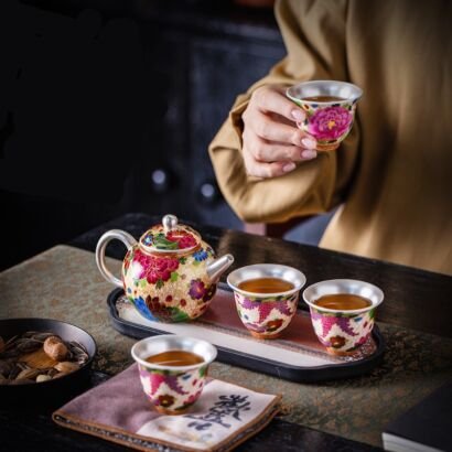 A person holding a small silver-lined ceramic teacup filled with tea, next to a matching floral cloisonne-style teapot and three small teacups set on a serving tray over a dark wooden tea table.