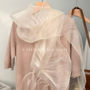 An off-white lightweight organza scarf with a silky sheen and delicate wavy feather-like ribbed textures, draped over a dusty pink knit top on a wooden hanger with fringe detailing at the bottom.
