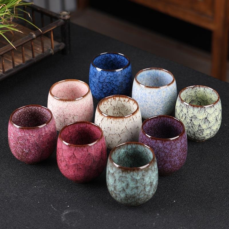 A cluster of small rounded ceramic teacups arranged on a dark surface, featuring rich kiln-transformed crackle glazes in cobalt blue, light blue, mint green, off-white, purple, magenta, pink, and turquoise with brown rims.