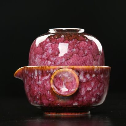 A compact handcrafted ceramic quick brewing teapot with an inverted matching teacup stacked on top, featuring a glossy magenta-crimson speckled glaze and a round heat-insulating side button against a dark background.