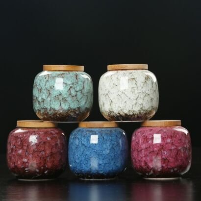 Five rounded handcrafted ceramic tea caddies topped with natural wooden lids, stacked in two tiers against a dark background, displaying rich kiln-transformed crackle glazes in turquoise green, off-white, deep red, royal blue, and magenta.