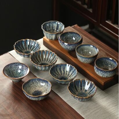 An array of handcrafted small ceramic teacups in various shapes—fluted, rounded, and petal-rimmed—arranged on a wooden tea table, showcasing deep ocean blue and cream speckled kiln-transformed glazes.